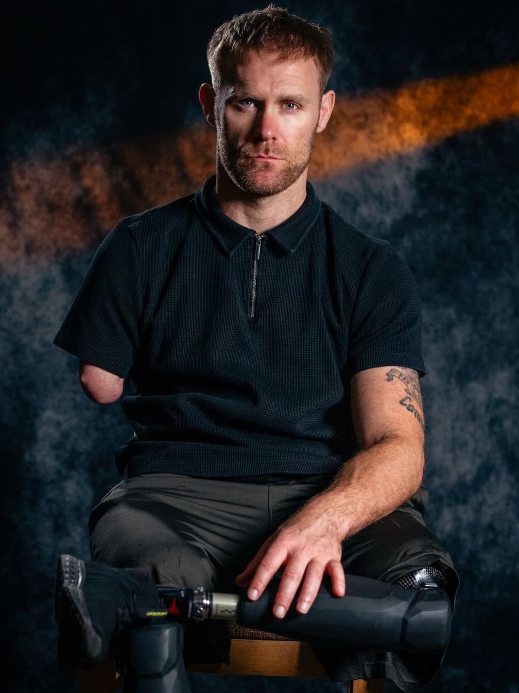 Mark Ormrod MBE, seated against a dark studio backdrop, prosthetic legs visible